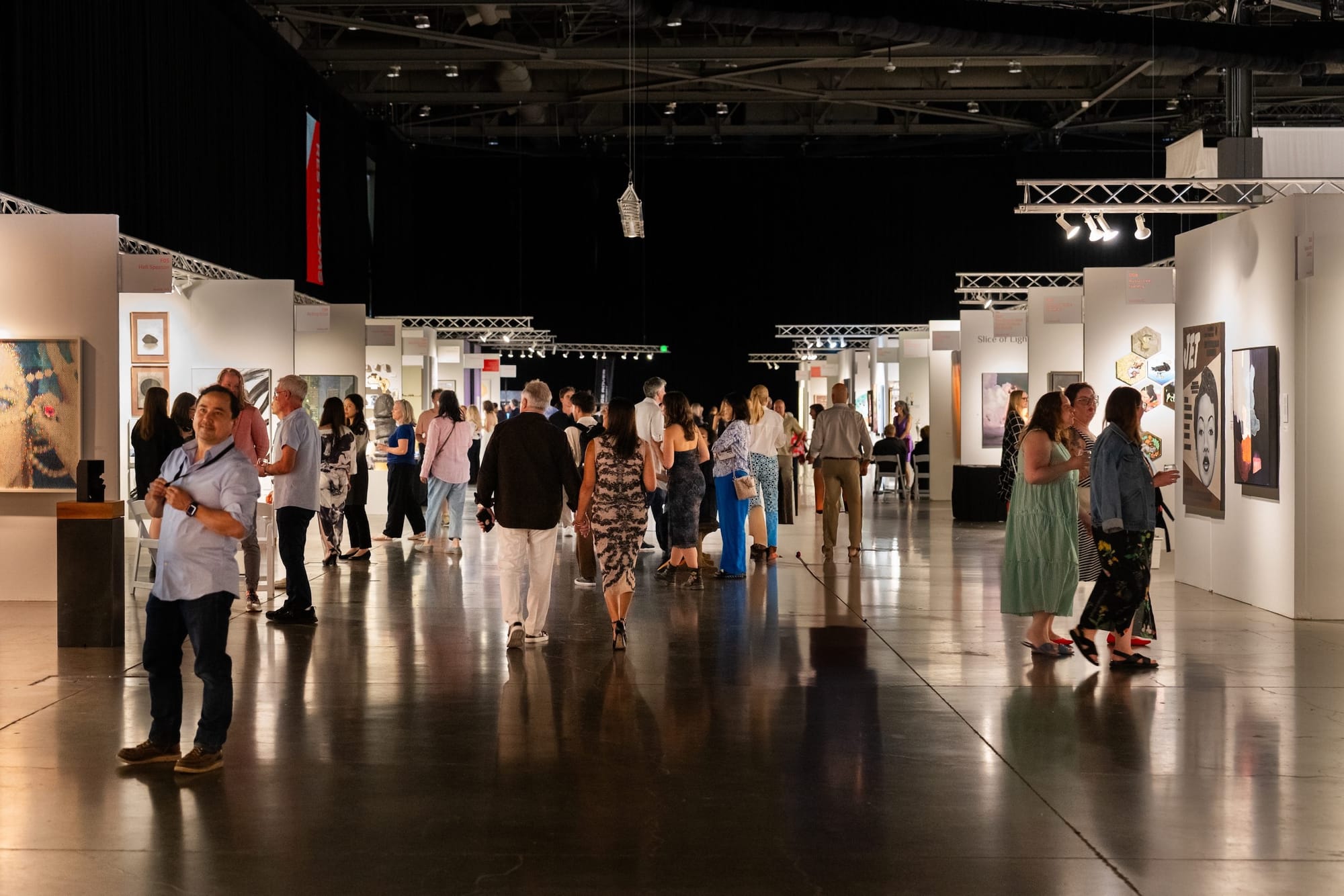 Photo of a crowd gathered inside white cubes on a convention floor, art hung on the cubes