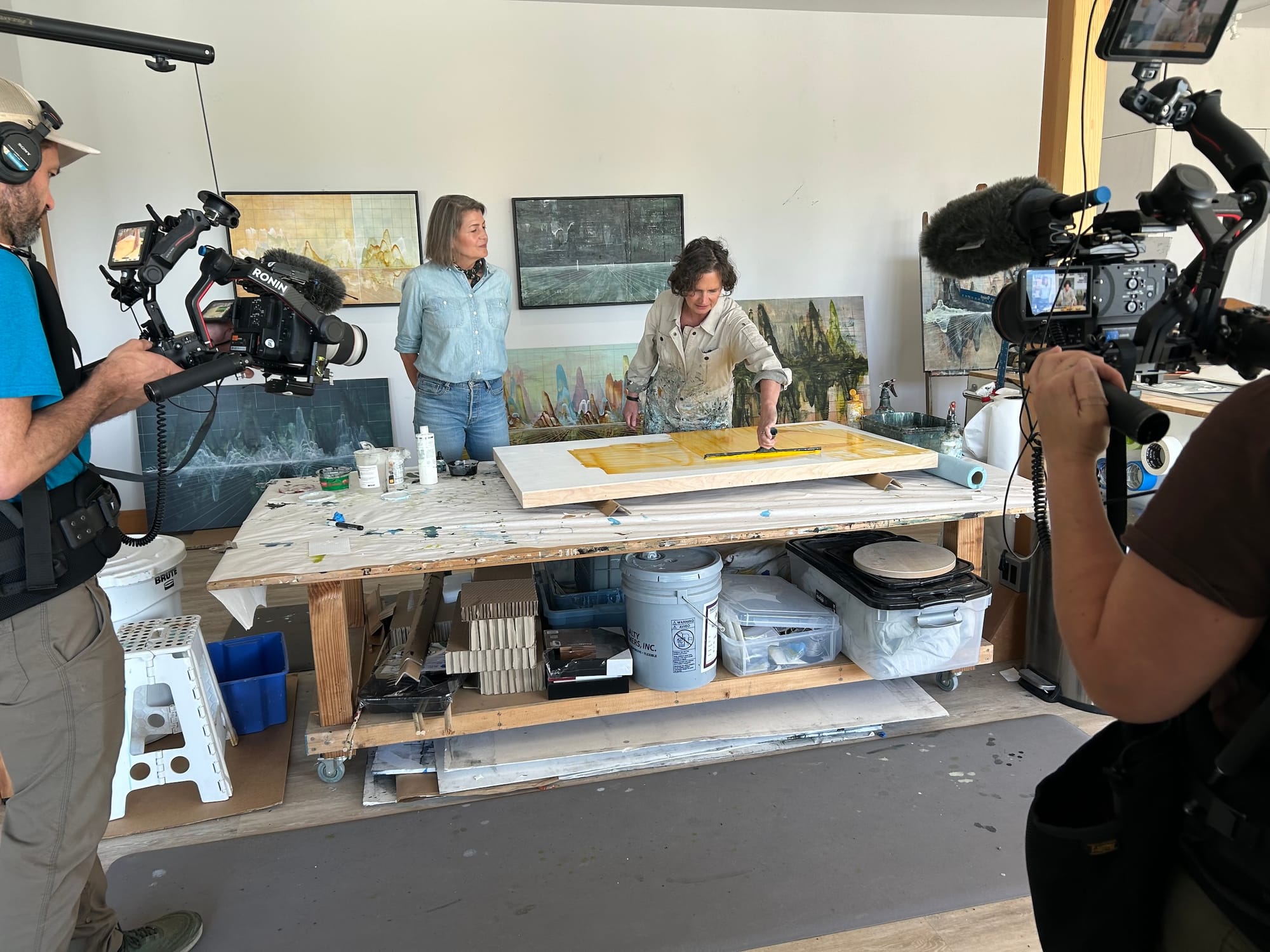 Photo of a film shoot in an art studio, with two people holding film cameras and two people at a painting table, with paintings hung behind