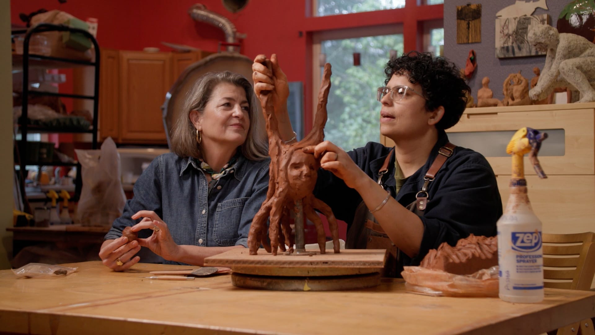 Two people in a clay studio -- one is watching as the artist makes a clay sculpture of a face with branches around it