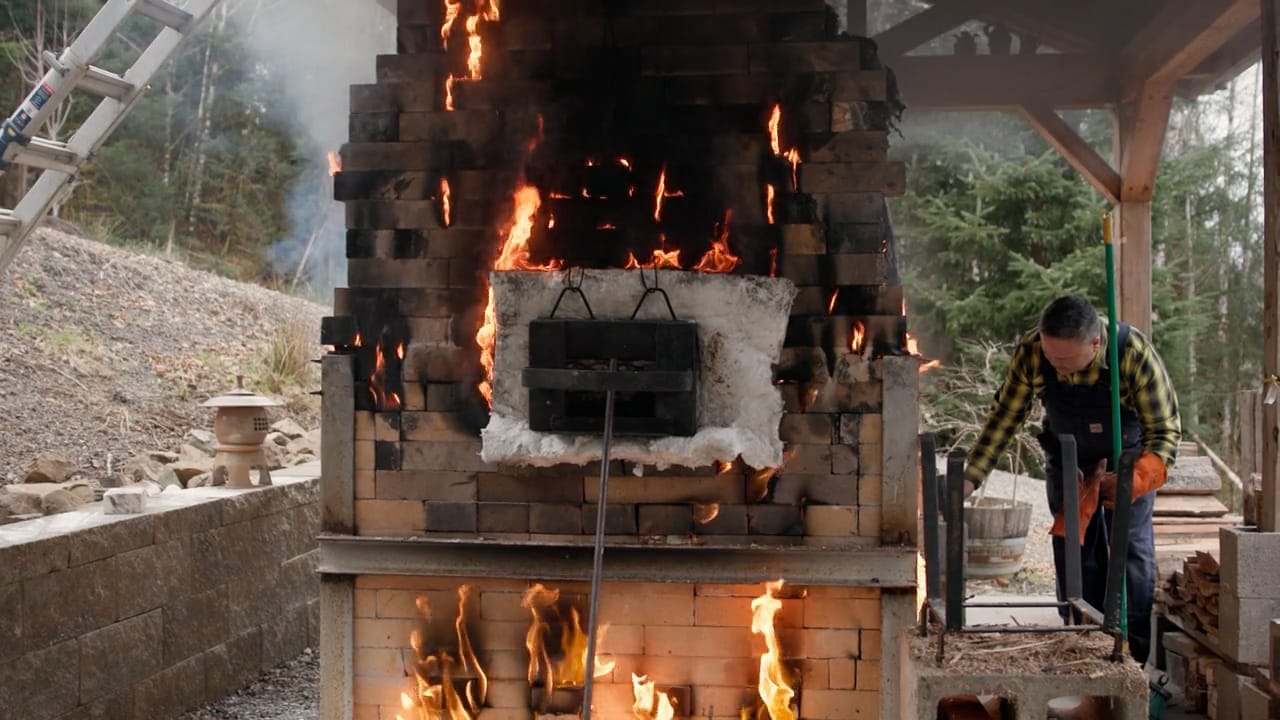 Photo of a man checking a tunnel-shaped kiln with flames licking out from the bricks.