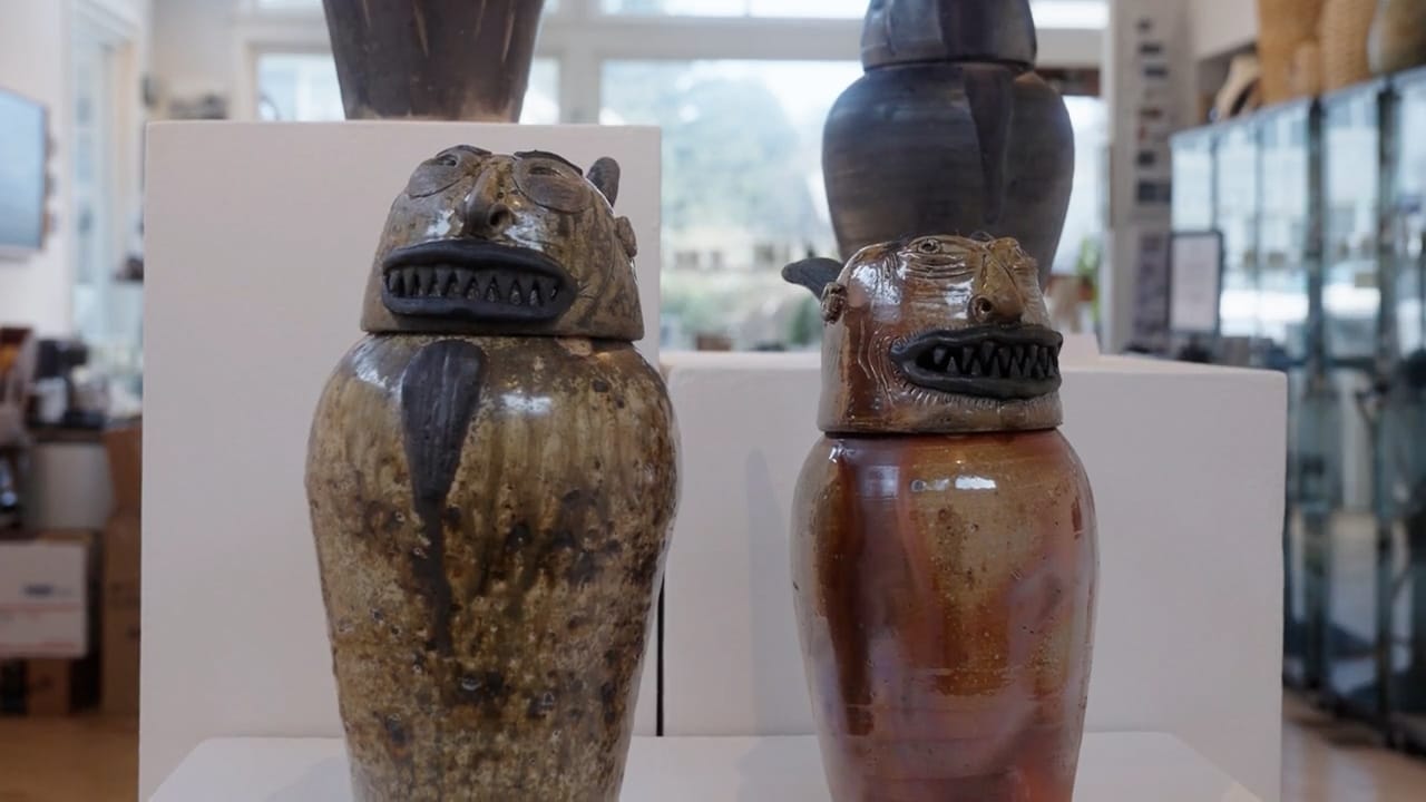 Photo of two pottery vessels at a gallery: lidded jars with monstrous faces and lots of teeth.