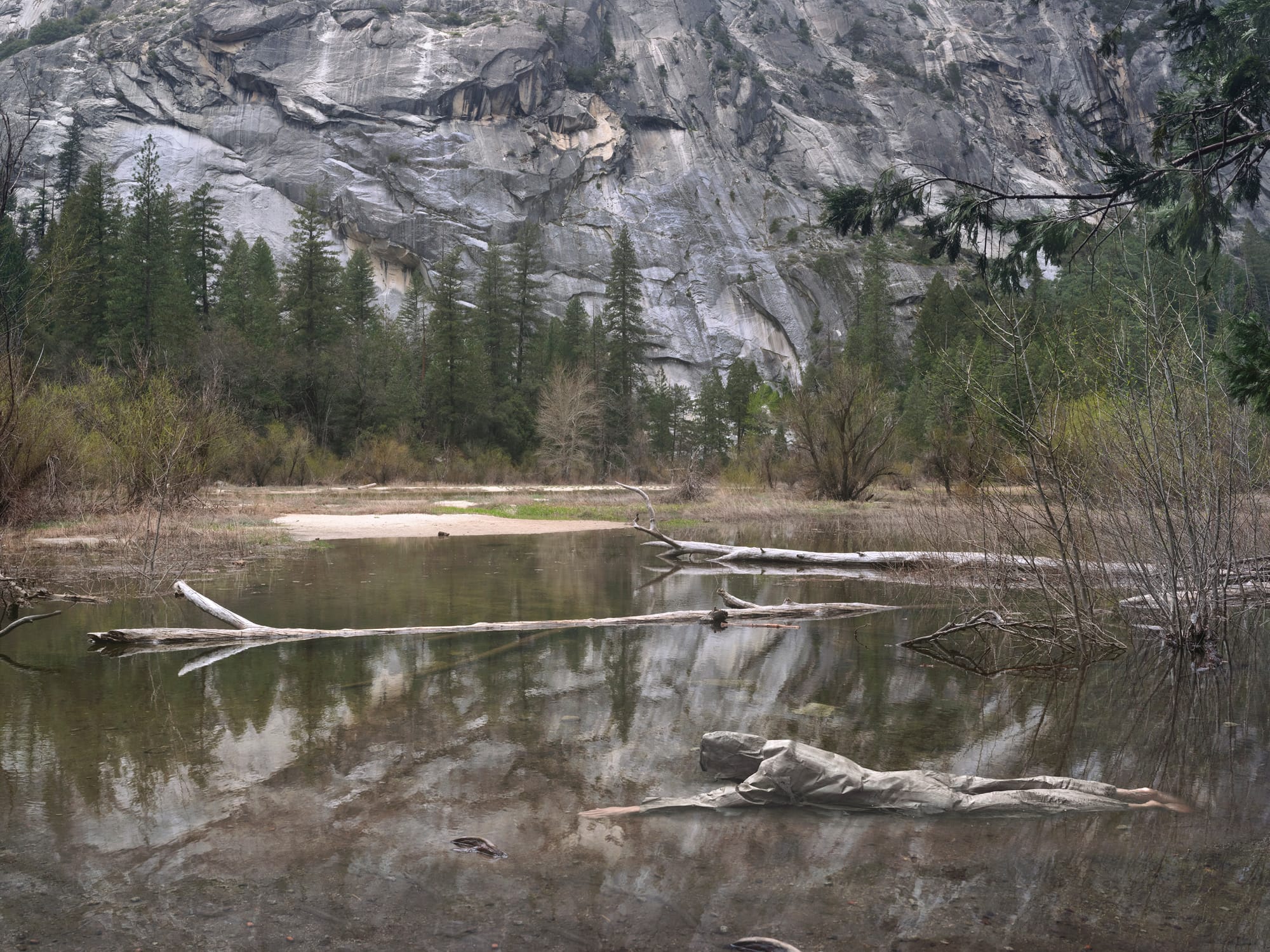Photograph of a natural landscape with rocky mountain slope and natural pool with dead logs and a person disguised as a dead log