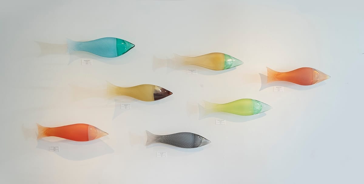 Photo of seven glass fish forms in bright colors hung on a wall, displayed as a school