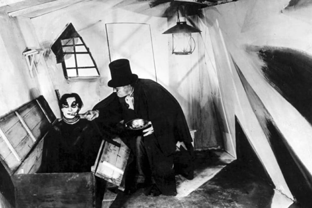 A black and white still from an old silent movie in which a top-hatted figure feeds a ghoul in a coffin.