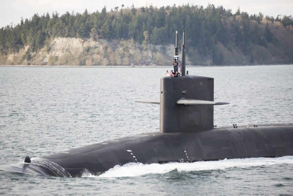 A submarine cruises across the surface of the water with a sailor on deck.