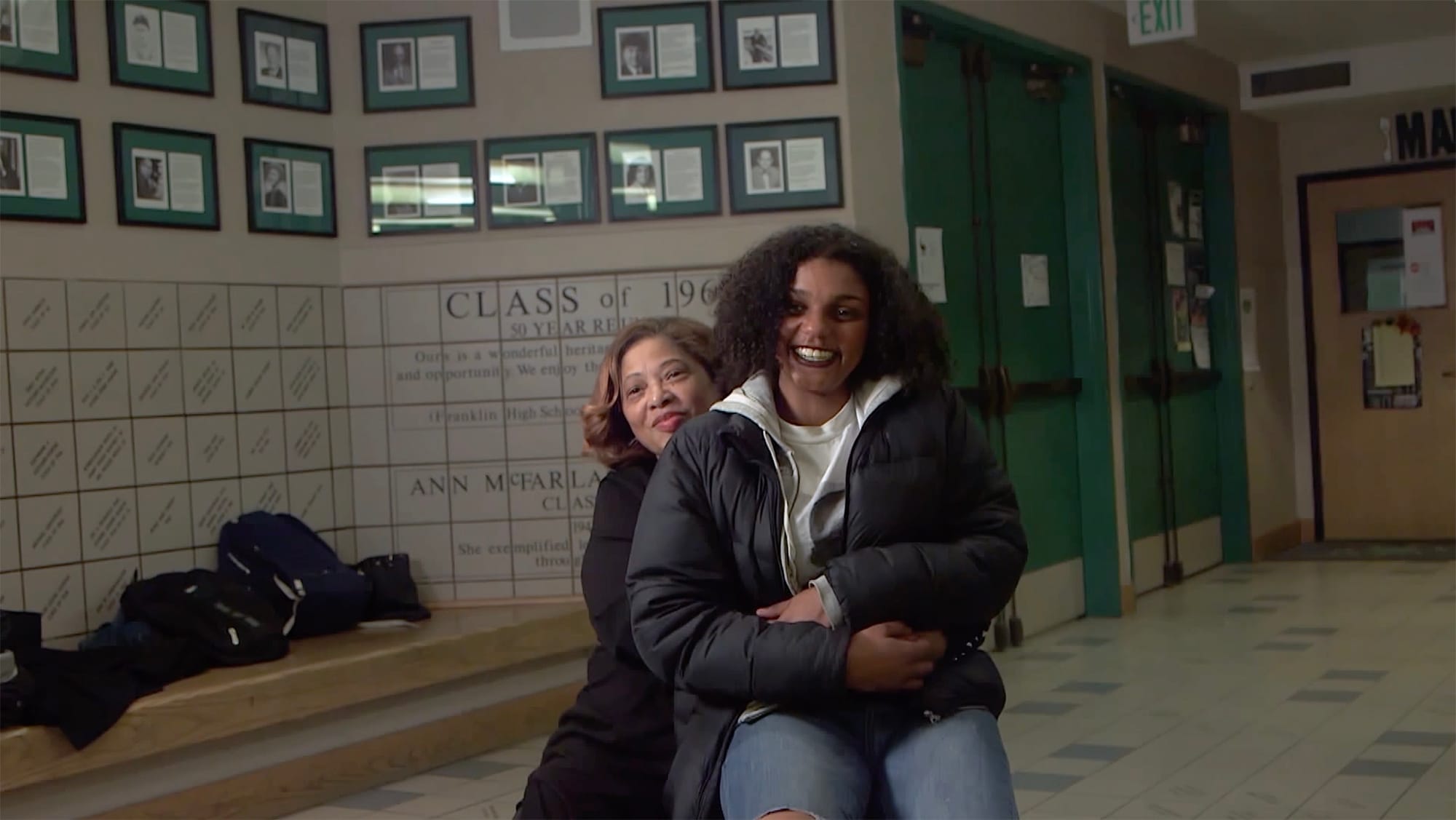 Woman, seated, with younger woman on her lap, in the hallway of a school.