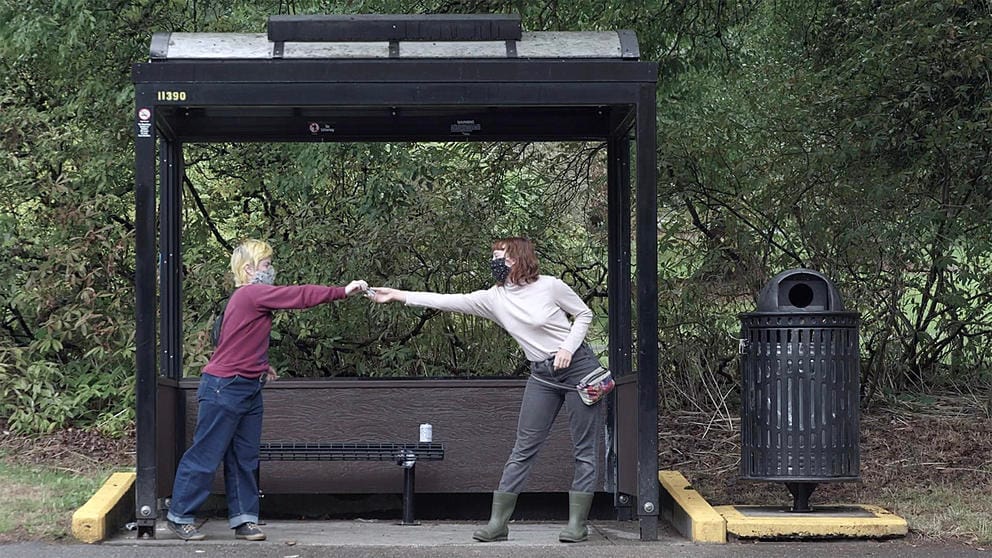 Two people in front of a bus stop, handing over hand sanitizer