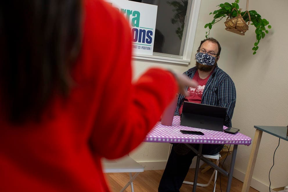 A woman with her back to the camera talks to a man seated in front of a laptop