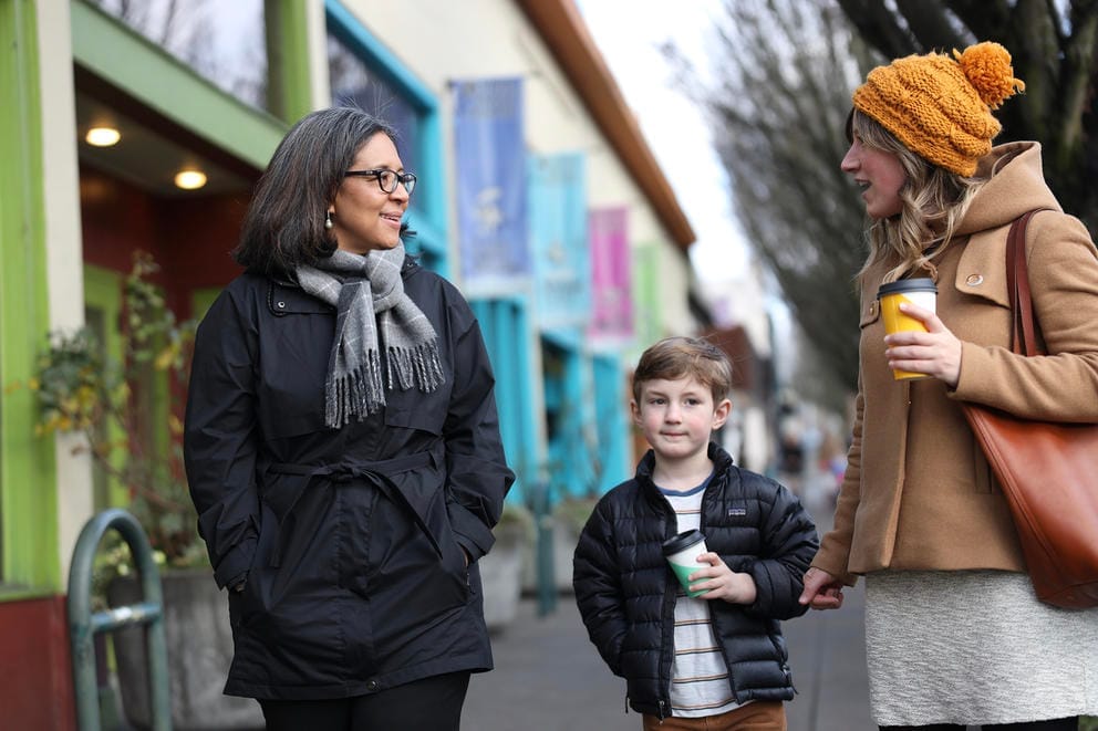 Marilyn Strickland talks to a woman and a child on a sidewalk, with colorful storefronts in background