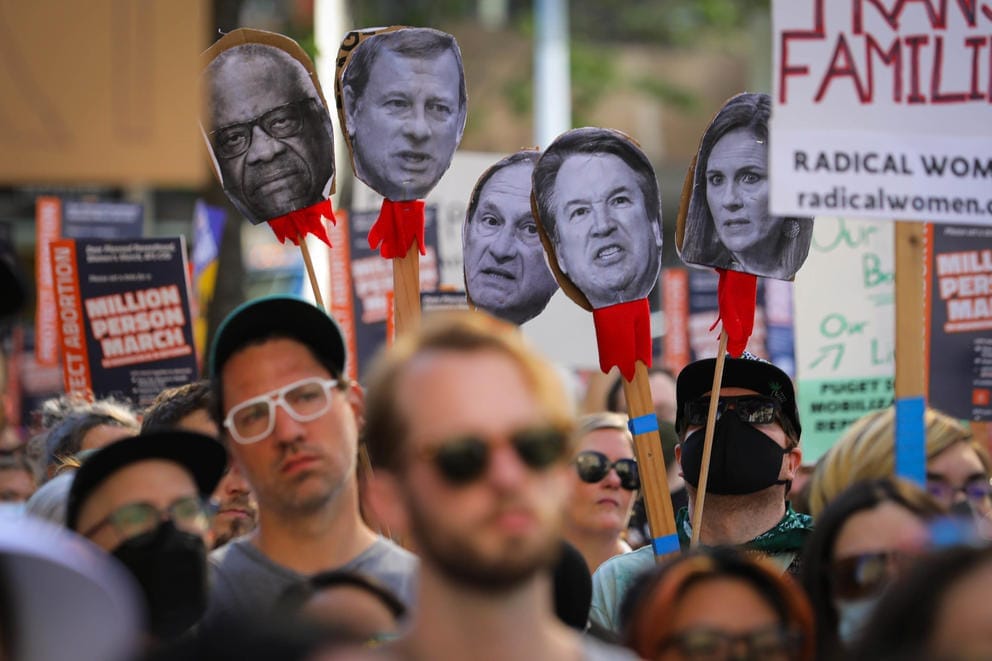 cutouts of five faces are held above a crowd of people