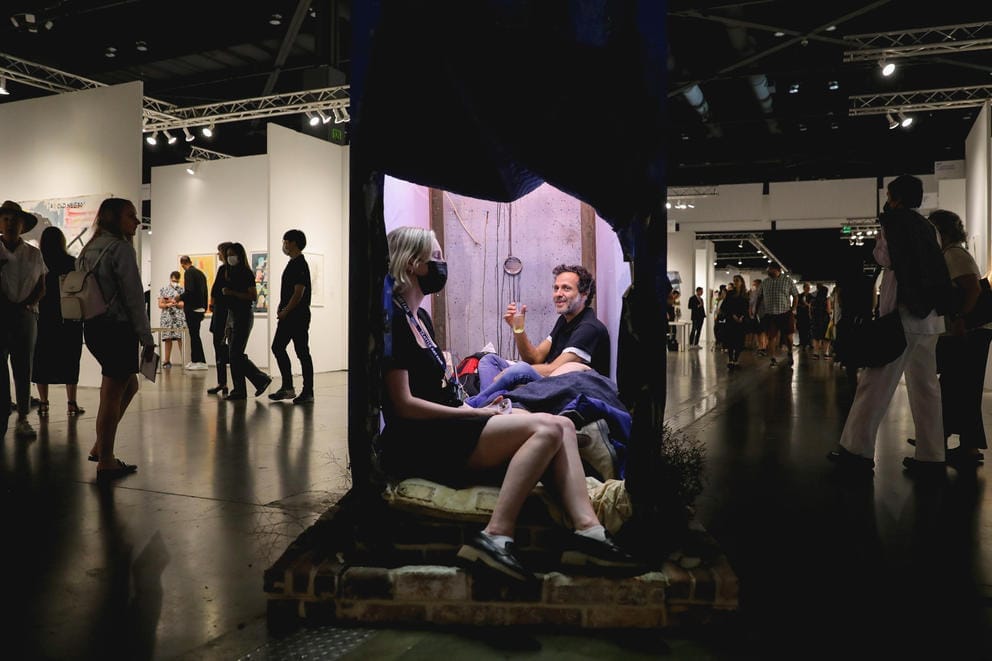 Two people sitting in a lit up small structure while art fair goes on around them
