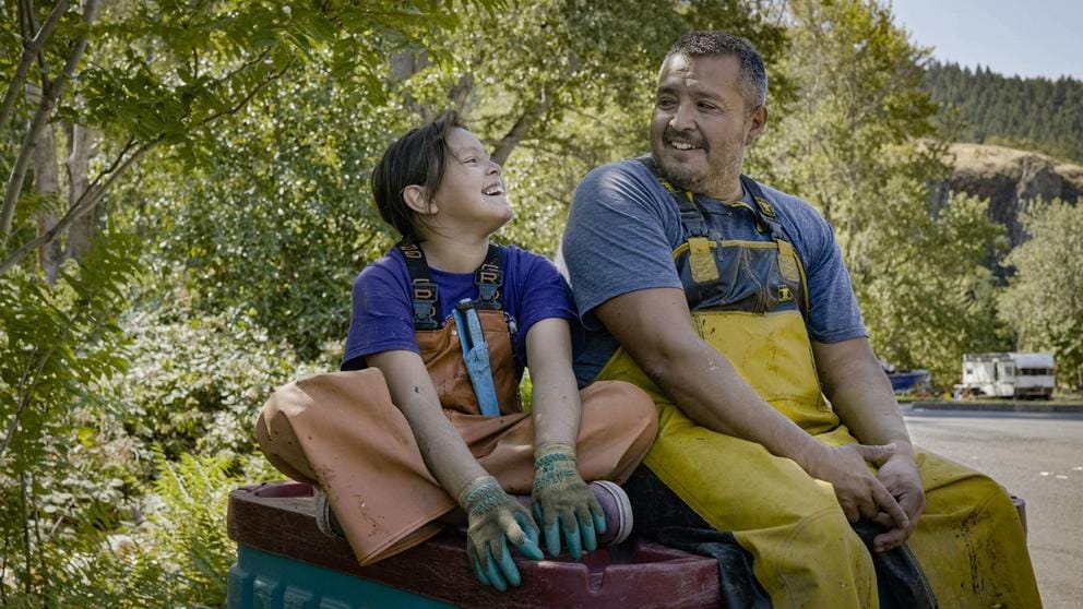 A young girl sits cross-legged while smiling up at her father sitting next to her. Both wear fishing waders.
