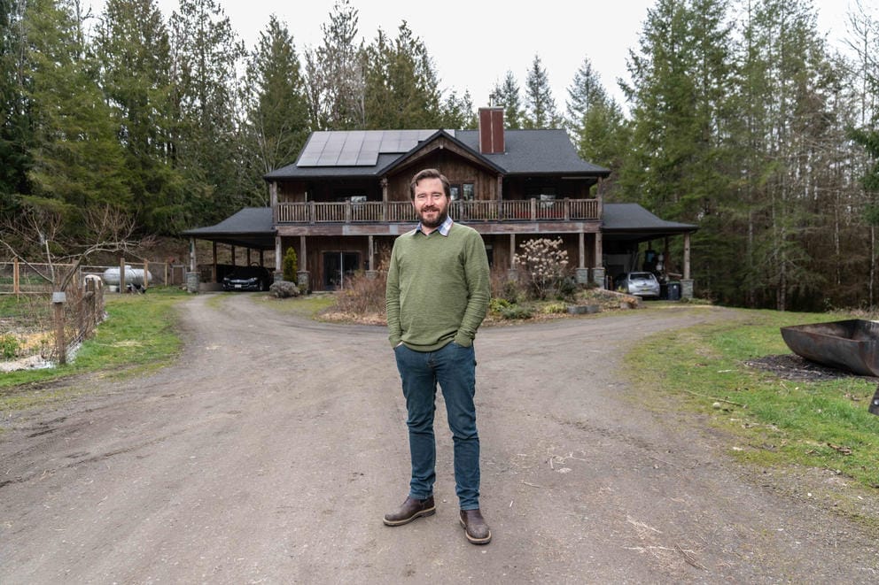 Greg Brotherton stands in a dirt driveway in front of his two-story house in a wooded area.