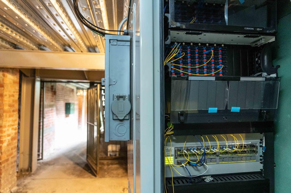 A gray panel box of fiber inputs and connections hangs on a wall in a tunnel.