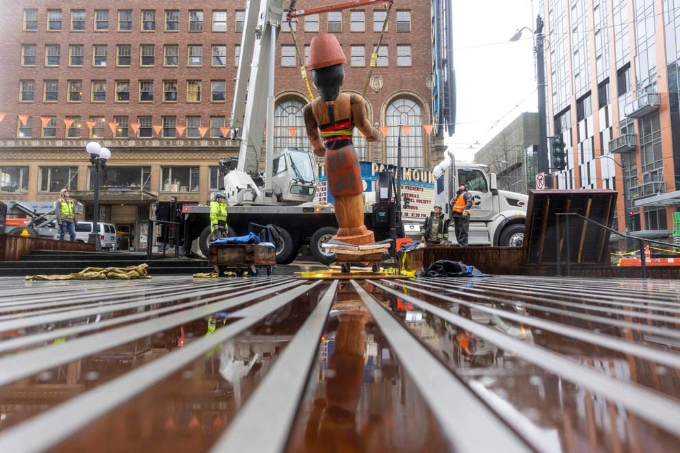 Workers stand outside using a crane to install “Mowitch Man,” one of two Salish welcome figures carved by Native American artist Andrea M. Wilbur-Sigo.