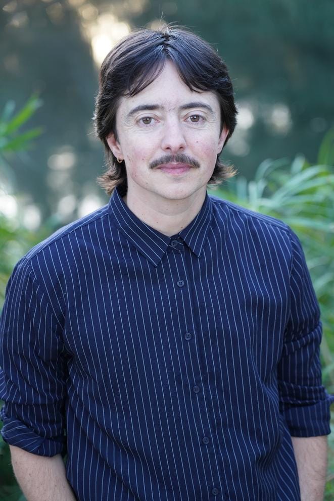Person in blue collared shirt with dark short hair and moustache looking at the camera