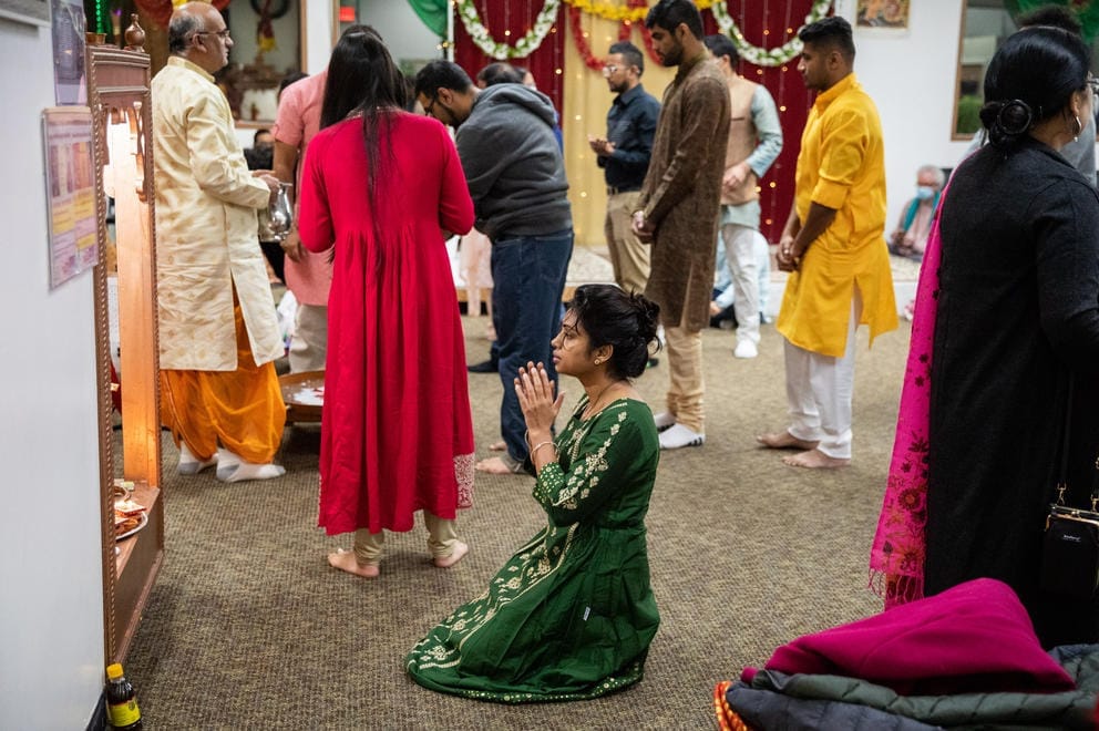 A woman in a green dress kneels in prayer. Other people in colorful outfits walk around behind her.