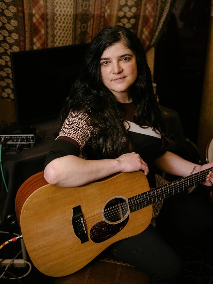 Person dressed in black facing the camera, resting arm on an acoustic guitar.