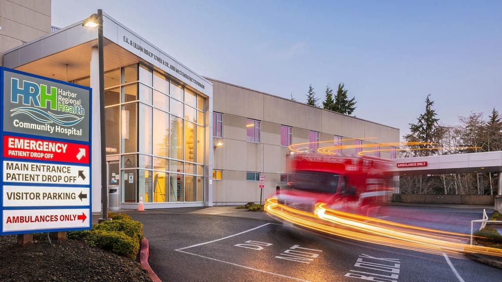 A long-exposure photo depicts an ambulance streaking past the front windows of the Harbor Regional Health emergency department.