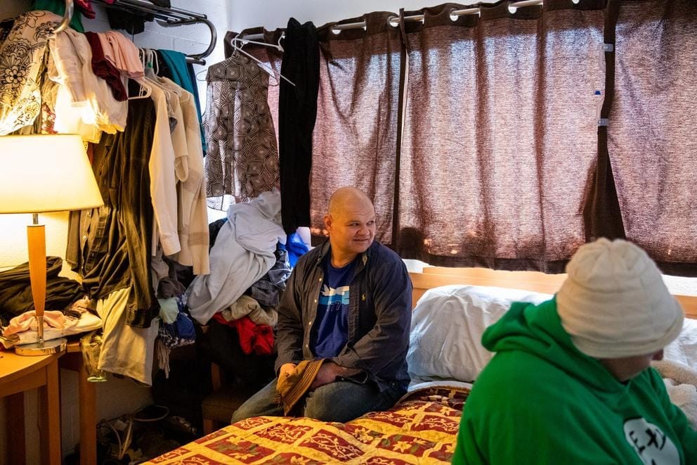 Man sits on a bed watching TV that is not in the picture, looking over his left shoulder. A woman sits on the other side of the bed looking at the TV that is to the right of the picture frame. Clothes hang in the background at left