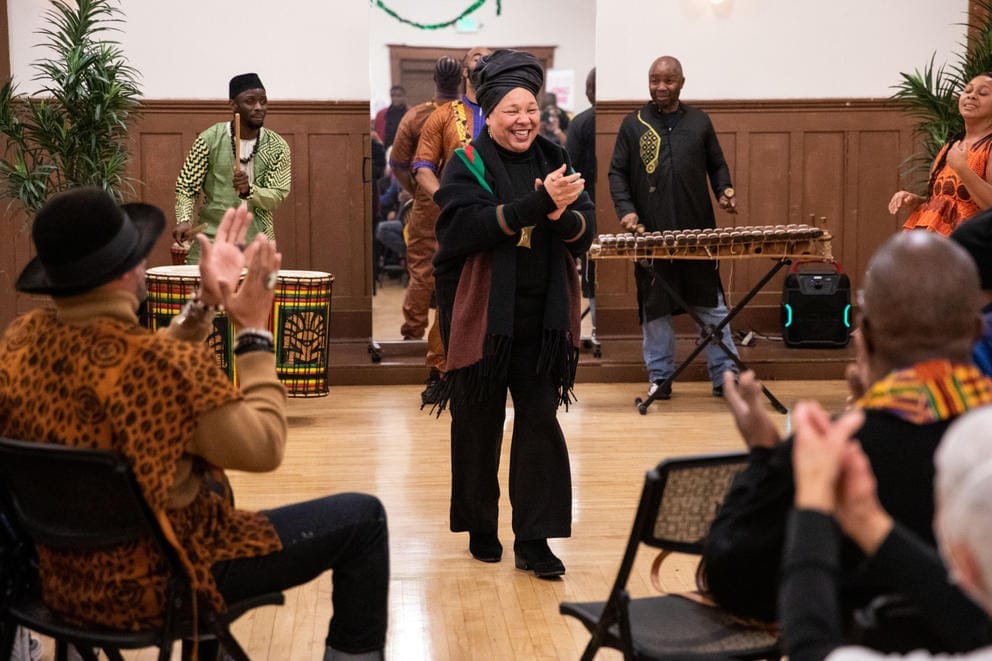 A woman, flanked by drummers, speaks to a crowd of seated people