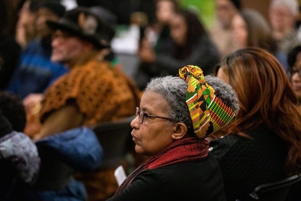 An older woman sits in a crowd, her hair is wrapped in a colorful kente cloth.
