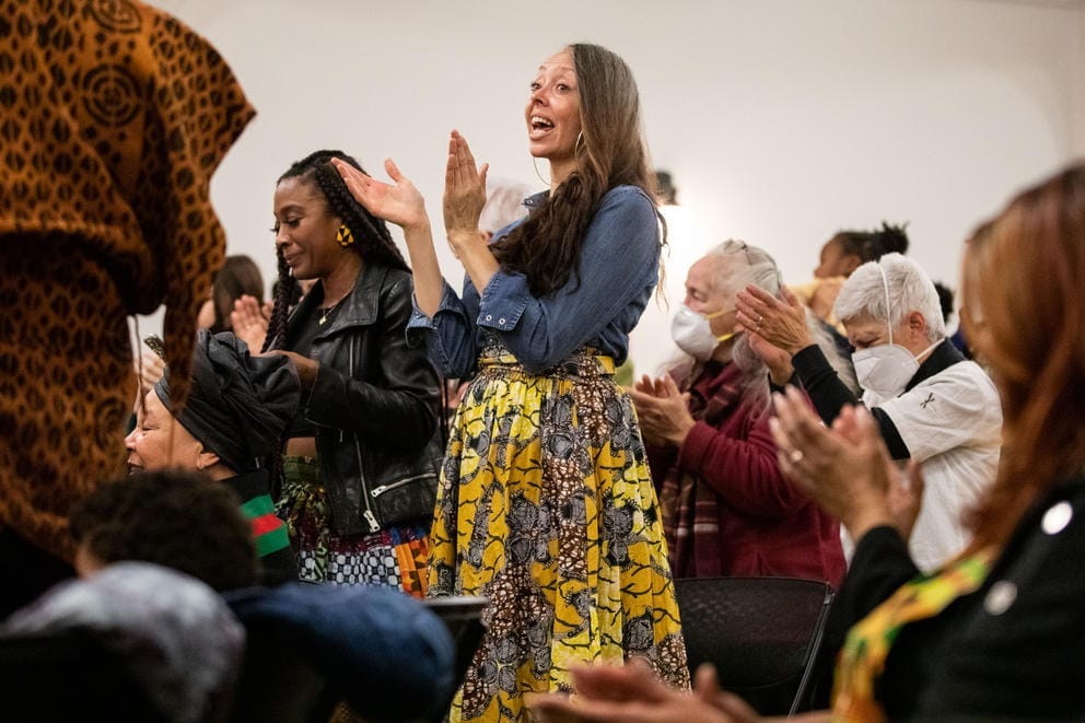 A mostly seated crowd applauds, a woman at center stands