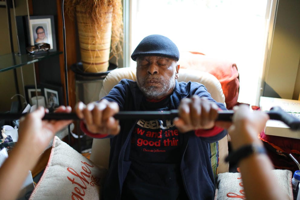 A man grips a cane held horizontally with both hands, another set of hands holds it up from out of frame