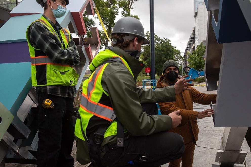 Two people in reflective vests and one person in a tan coat working on a metal sculpture on the right