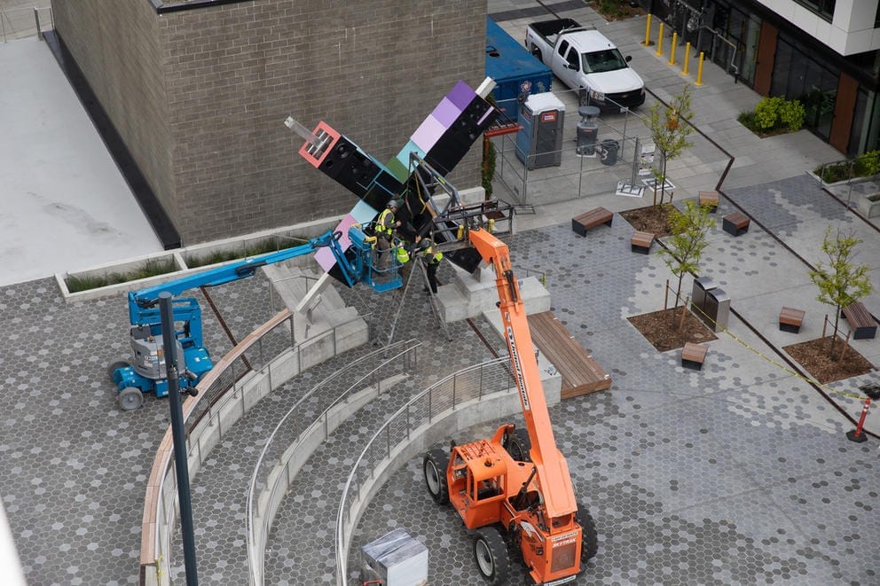 Orange forklift on gray tiled plaza near an X shaped sculpture made out of boxes that are made to look like speakers and have differently hued sides: pink, blue and purple.