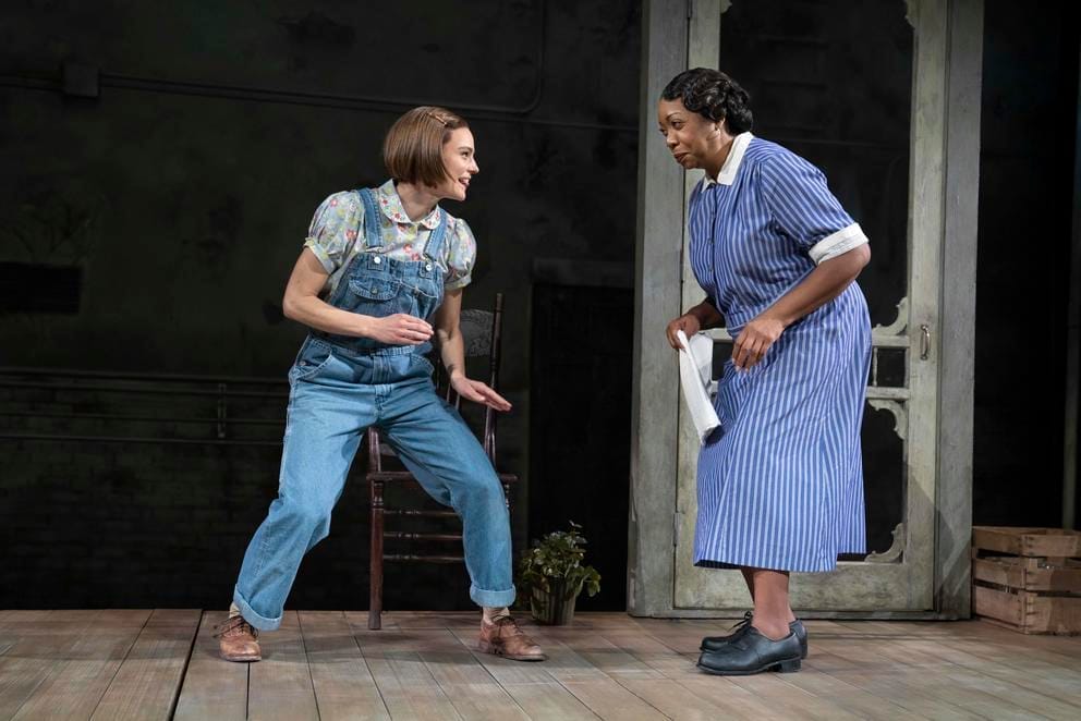 A young white girl in overalls stands to the left, to the right an older Black woman in a blue dress