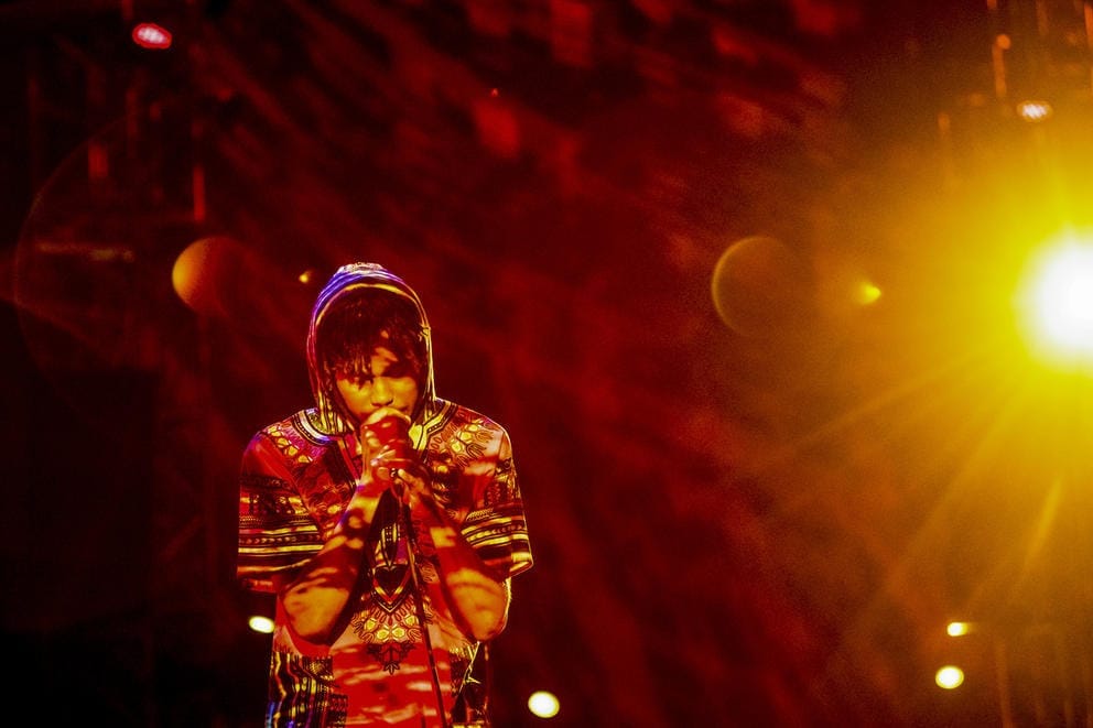 Person in shirt with African fabric, wearing a surgical mask and singing into a microphone. the light is orange and red, and a light source shines into the camera from the right side of the photo.