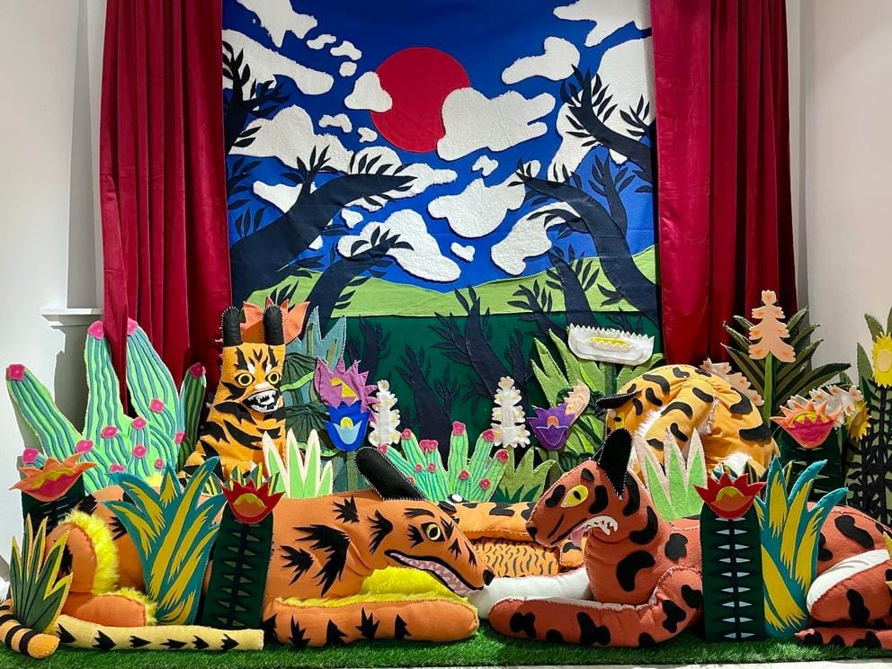 Animal puppets sit within exotic plants in front of a blue sky background with clouds — all made out of felt.