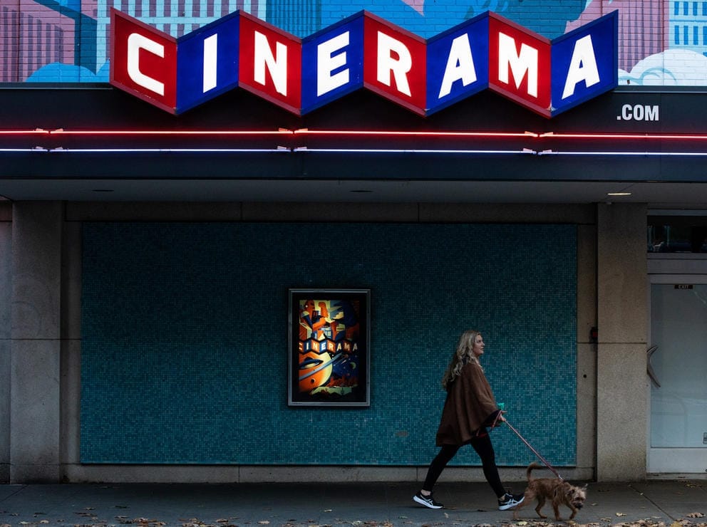 Woman walks her dog in front of a dark facade, up top red and blue squares with white letters spell out CINERAMA
