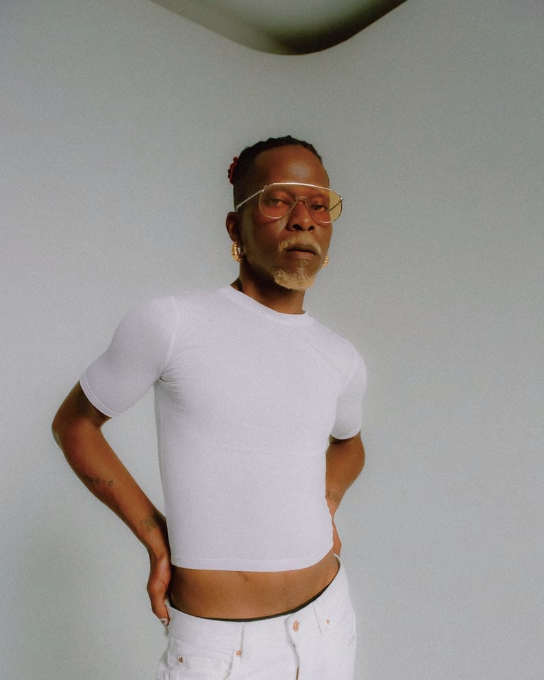 Pierre Kwenders looks at the camera in a white t-shirt, white pants and yellow-tinted sunglasses with a hand on his hip.