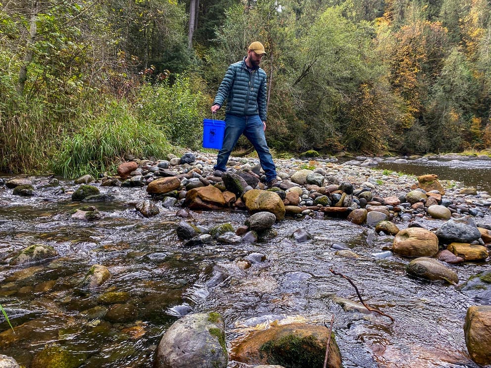 person holding bucket walks across rocks in a river