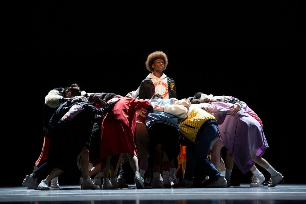 a photo of dancers performing on stage, one young man at the center of many others