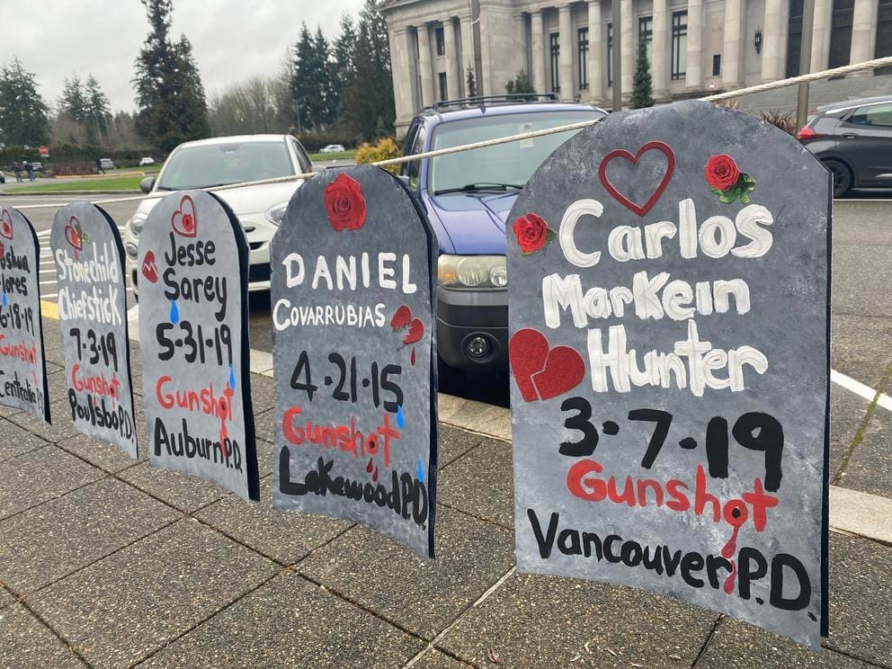 Four signs in the shape of gravestones bear the names of people killed by police and the dates they died. From right to left: Carlos MarKein Hunter, 3-17-19, Gunshot, Vancouver PD; Daniel Corrubias, 4-21-15, Gunshot, Lakewood PD; Jesse Sarey, 5-31-19, Gunshot, Auburn PD; Stonechild Chiefstick, 7-3-19, Gunshot, Poulsbo PD.