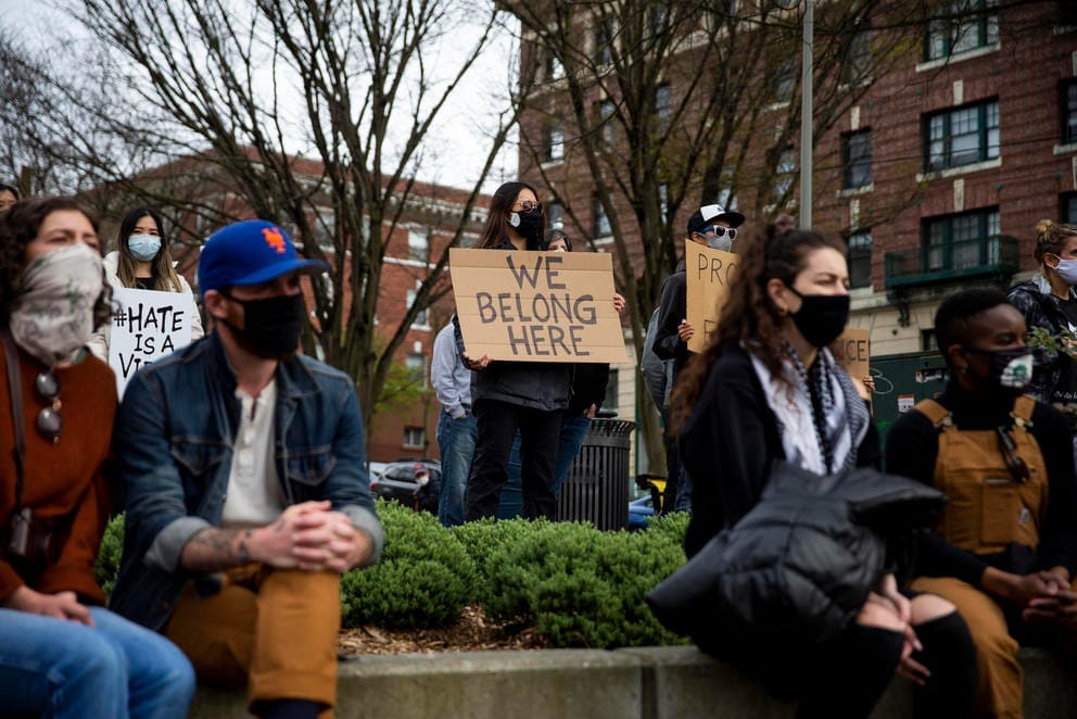 masked people gather at an outdoor rally, a central figure holds a sign that reads