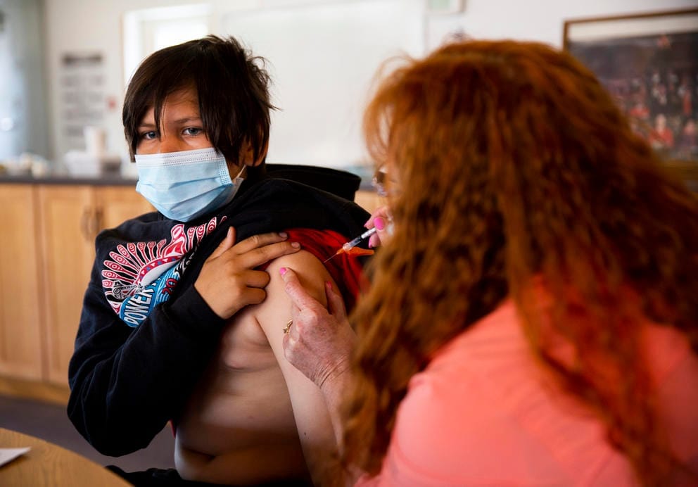Barbara Hoffman administers a vaccine to Urijah Woodward who is wearing a mask and has his sweatshirt pulled up over his shoulder