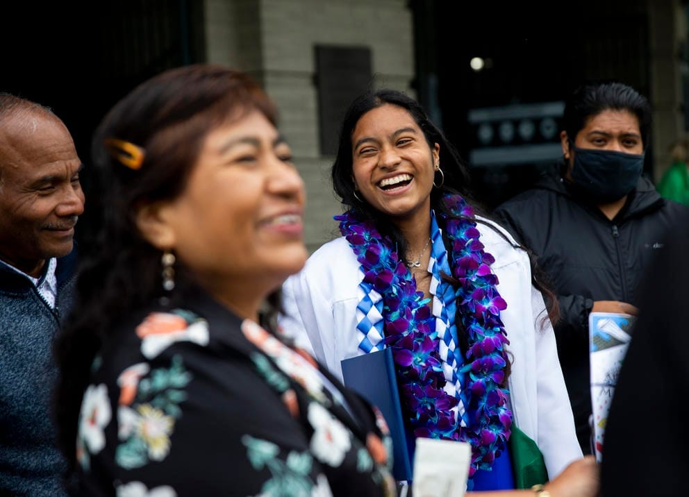 Donaji Torres-Marquez, wearing a white graduation gown and blue and purple leis, smiles surrounded by family members