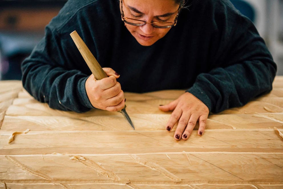 Andrea Wilbur-Sigo is bent over a large cedar log with a chisel in her hand carving designs into the wood