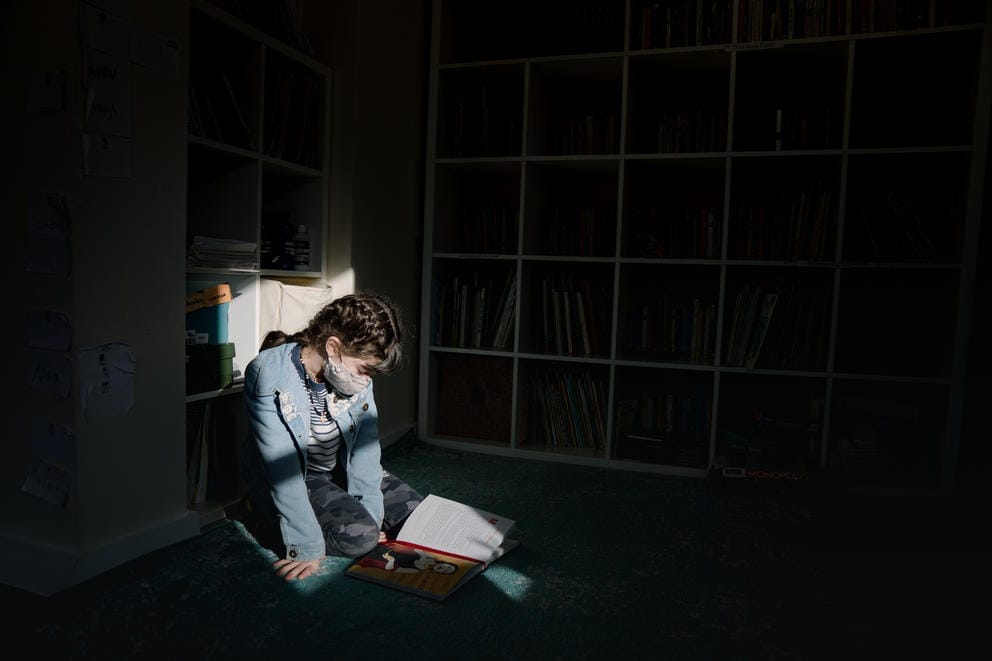 A girl is illuminated by a patch of window light, she is wearing a mask, sitting on the floor and looking down at a book