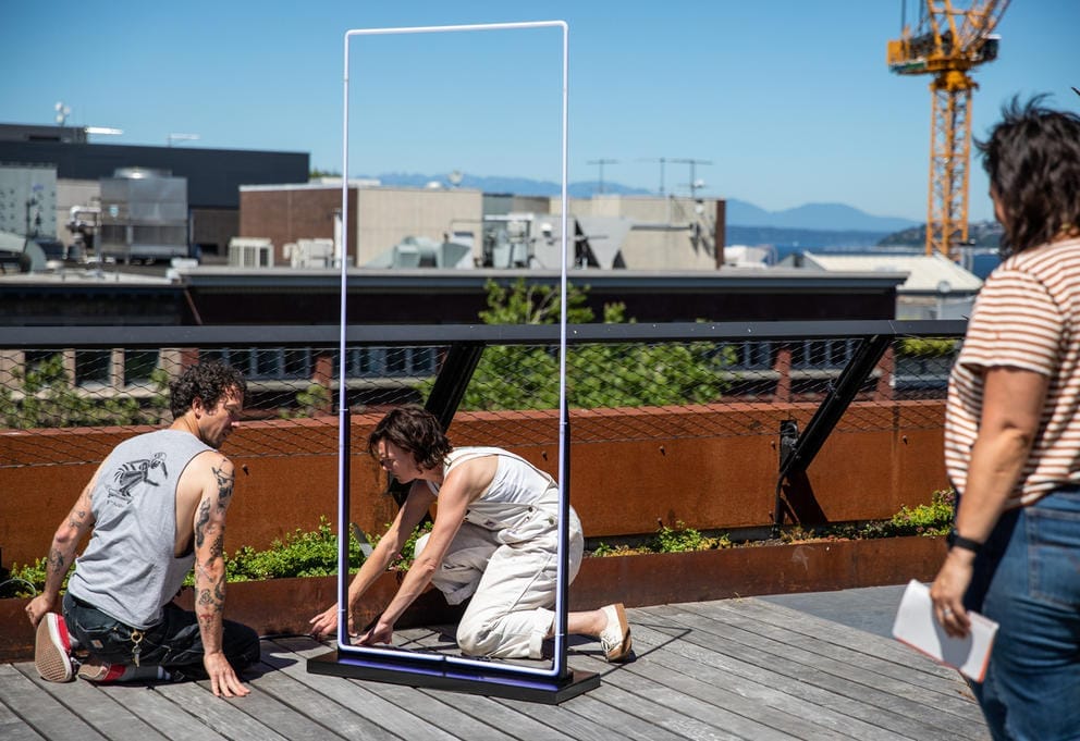 three people on a balcony with views of the water and city work on an installation, which is in the shape of a tall rectangle made out of neon