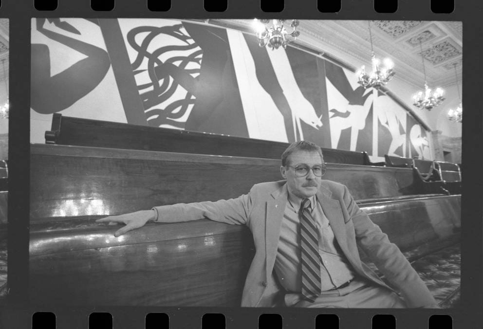 black and white photo of a man in a suit sitting on a bench in front of a large mural