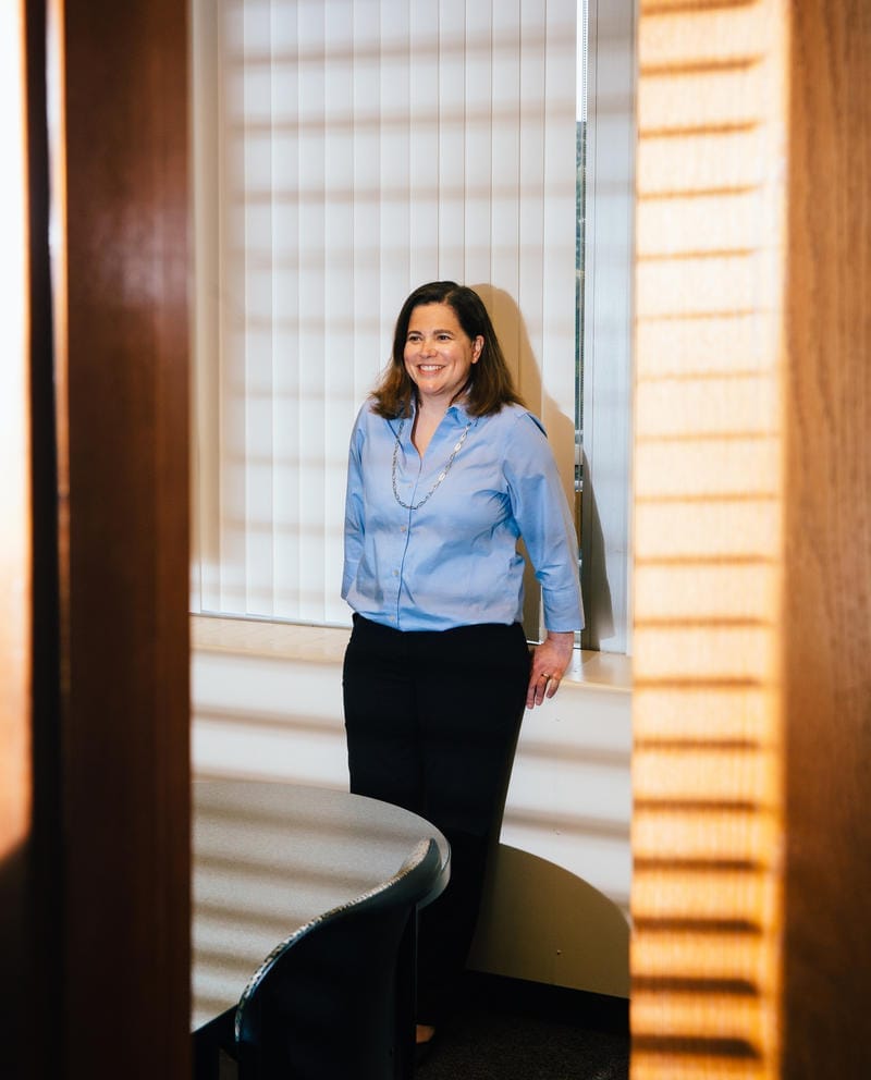 Sadie Armijo stands against a wall at the State Auditor's Office as sunlight coming through the blinds stripes across her and the room.