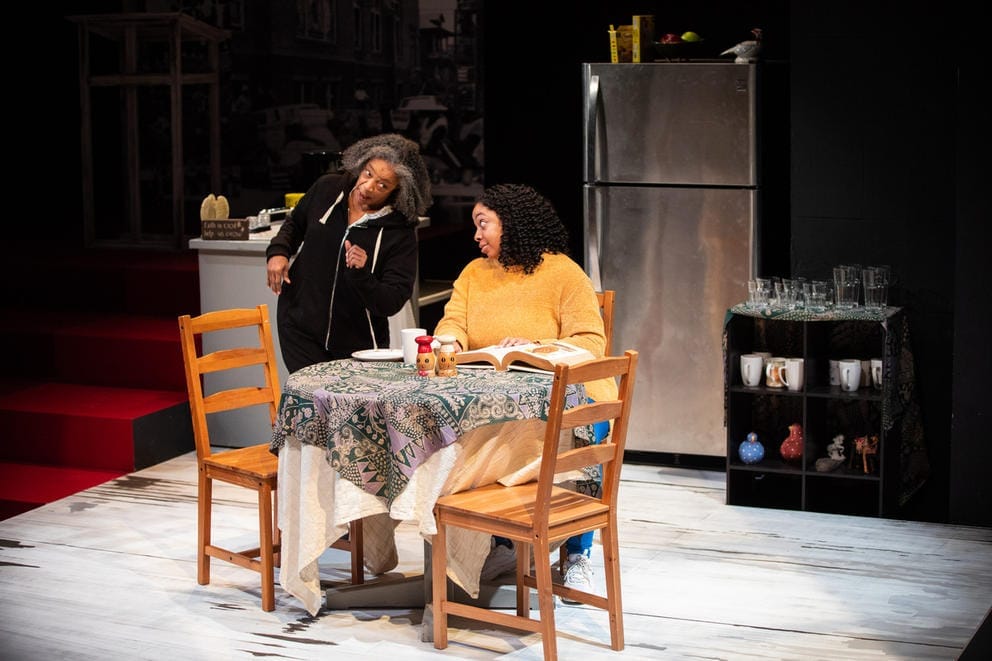 Two people on a stage, one person sits at table and another stands nearby
