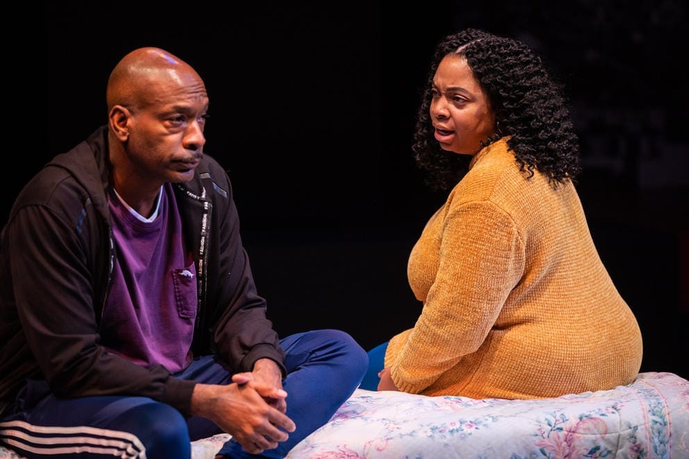 Two people sit on a bed, a man on the left and woman on the right.
