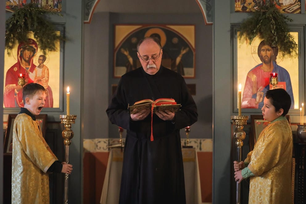 Two altar boys stand on either side of a clergy member reading from a Bible. The boy on the left yawns.