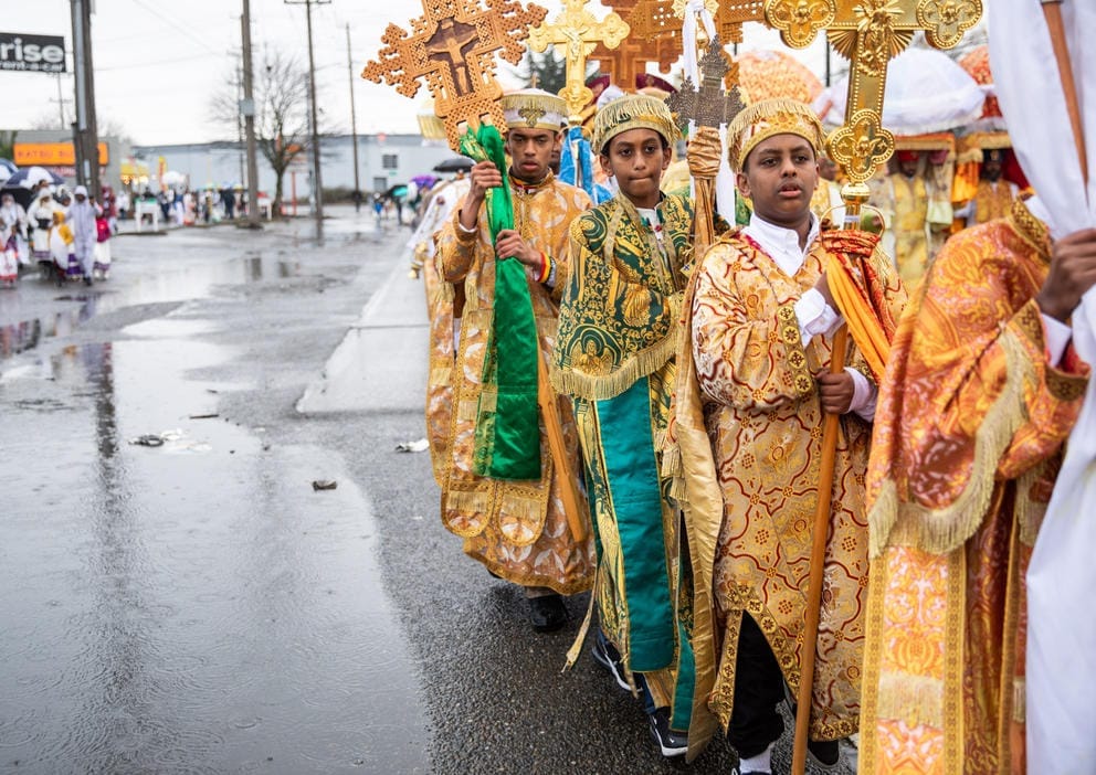 Altar boys in gold robes and traditional garb hold crosses outside in the rain